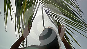 The girl goes through a branch of a palm tree to the sea resting on the beach and the sun shines through the palm tree