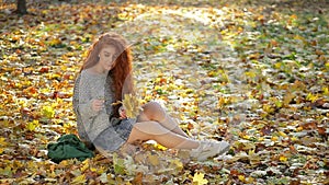 Girl with gorgeous thick curly red hair sits in beam of contour light on blanket of yellow autumn leaves and collects bouquet