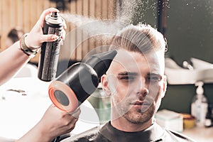 Girl hairdresser sprays spray and dries hair with a hairdryer to a young guy sitting in a chair in a barbershop