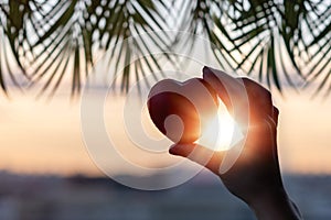 Girl hand holding red heart against the background of the sea in branches of palm trees. Sunset beach. Summer and freedom concept.