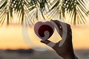 Girl hand holding red heart against the background of the sea in branches of palm trees. Sunset beach. Summer and freedom concept.
