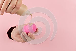 Girl hand holds a beauty blender in foundation, concealer from a hole in a pink background. Makeup artist concept, copy space