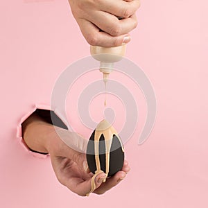 Girl hand holds a beauty blender in foundation, concealer from a hole in a pink background. Makeup artist concept, copy space