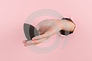 Girl hand holds a beauty blender for makeup from a hole in a pink background, copy space