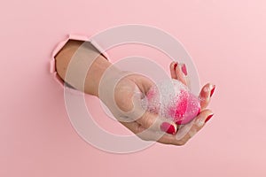 Girl hand holds a beauty blender for makeup from a hole in a pink background, copy space. Sponge in soapy foam, how to clean