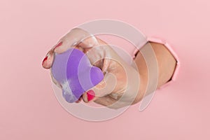 Girl hand holds a beauty blender for makeup from a hole in a pink background, copy space. Sponge in soapy foam, how to clean