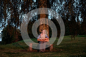 Girl with a harp sits near a birch at sunset