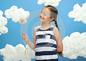 Girl has a big dandelion in her hands, dressed in striped dress, posing on a blue background with cotton clouds, the concept of su