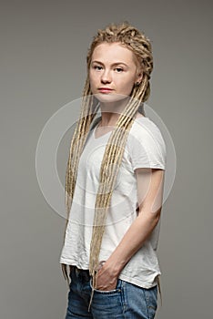 Girl has long dreadlocks, blonde hair, standing on gray background, hands in pockets