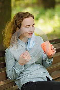 A girl has removed her surgical mask to take a sip from a mug.