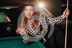 A girl in a hat in a billiard club with a cue and balls in her hands.Playing pool