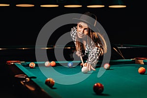 A girl in a hat in a billiard club with a cue in her hands hits a ball.Playing billiards