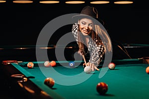 A girl in a hat in a billiard club with a cue in her hands hits a ball.Playing billiards