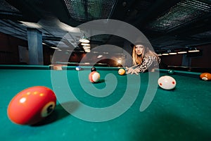 A girl in a hat in a billiard club with a cue in her hands hits a ball.Playing pool