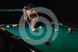 A girl in a hat in a billiard club with a cue in her hands hits a ball.Playing pool