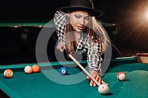 A girl in a hat in a billiard club with a cue in her hands hits a ball.Playing pool