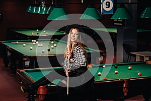 A girl in a hat in a billiard club sits on a billiard table with a cue in her hands.Playing billiards