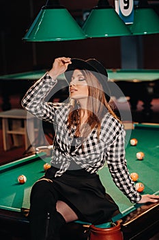 A girl in a hat in a billiard club sits on a billiard table.Playing pool