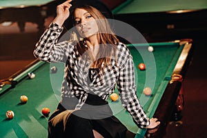 A girl in a hat in a billiard club sits on a billiard table.Playing pool