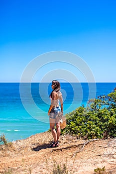 A girl with a hat in her hands is standing with her back on a rock against the blue ocean