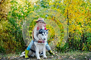 Girl in hat hugging her dog. Little girl with her dog, alaskan malamute, full length. Child and dog on nature background