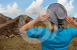 Girl in hat looks at mountain terrain and thick clouds and blue sky from a cliff. White middle aged woman stays back to us and