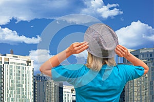 Girl in hat looks at on thick clouds and blue sky with skyscrapers or urban buildings on background. White middle aged woman stays