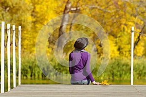Girl in a hat sits on the dock and admires the colors of autumn.