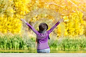 Girl in a hat sits on the dock with her hands up. Back view