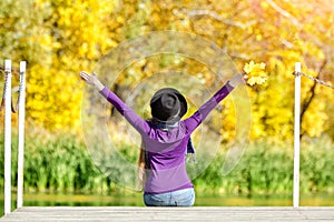 Girl in a hat sits on the dock with her hands up. Back view