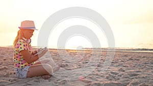 A girl in a hat sits on the sand and works on a tablet PC