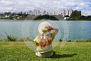 Girl with hat sitting on grass in Barigui Park enjoying Curitiba cityscape, Brazil photo