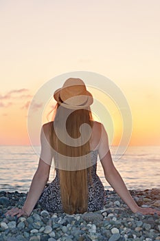 Girl in the hat sitting on the seashore. Sunset time. Back view
