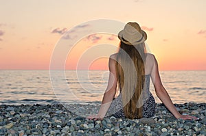 Girl in the hat sitting on the seashore. Sunset time. View from the back