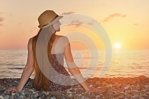 Girl in the hat sitting on the seashore. Sunset time. View from the back