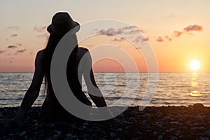 Girl in the hat sitting on the seashore. Sunset time. View from the back, silhouettes