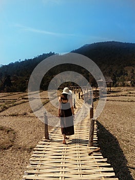 A Girl in the hat walking to bamboo bridge in rice fields on the background of the mountain