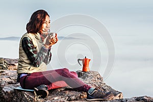Girl is having breakfast and drinking coffee on a cliff top with a view of sunrise in mountains, and a thick fog is creeping