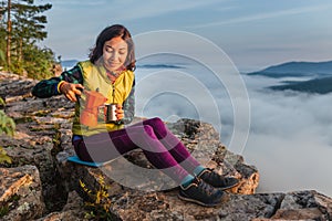 Girl is having breakfast and drinking coffee on a cliff top with a view of sunrise in mountains, and a thick fog is creeping