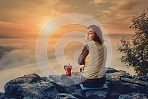 Girl is having breakfast and drinking coffee on a cliff top with a view of sunrise in mountains, and a thick fog is creeping