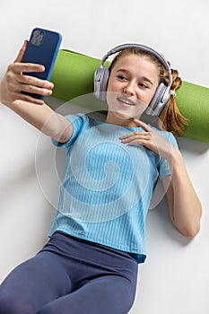 Girl in headphones with a phone, is resting after yoga. Green rug roll under your head.