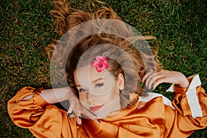 A girl with healthy beautiful hair lies on the grass and looks at the camera. Girl with perfect skin and natural care. Flower in