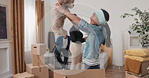 Girl helps parents unpack boxes after moving finds cardboard box with her toys pulls out teddy bear is happy hugs it mom