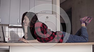 A girl in her 20`s lying on kitchen table surface and typing something in laptop.