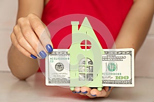 The girl in her hands holds a small green house and one hundred dollar bills, money