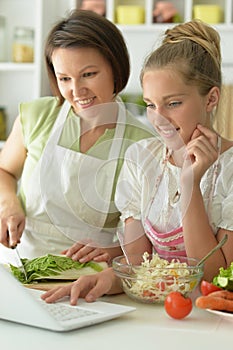 Girl with her mother cooking together at kitchen table looking at laptop