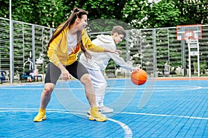 Girl and her younger brother, teenager, play basketball on modern basketball court under open sky