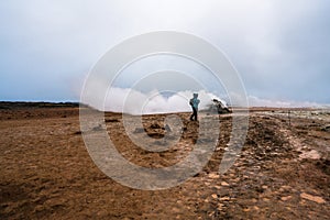 Girl with hiking gear infront of sulphur steam vents in Iceland during heavy cold wind. Mineral rich and textured muddy ground inf