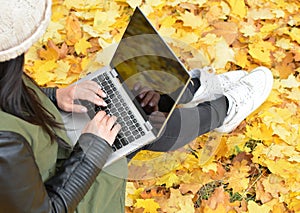 Girl in hipster with laptop in autumn park. A woman in a cap using a laptop while sitting on fallen leaves. Freelancer in the hat