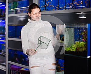 Girl holding aquarian net and water container next to aquarium with colorful fish in aquarium shop